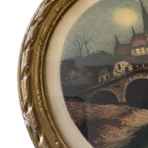 Puede incluir: Una impresi&oacute;n antigua enmarcada de forma ovalada. La impresi&oacute;n representa un paisaje nocturno con un puente, edificios y una luna llena. El marco es de color dorado con un dise&ntilde;o detallado y ornamentado. La obra de arte es vintage.