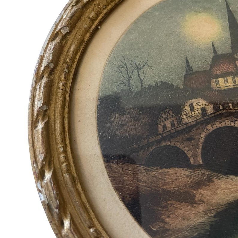 Puede incluir: Una impresi&oacute;n antigua enmarcada de forma ovalada. La impresi&oacute;n representa un paisaje nocturno con un puente, edificios y una luna llena. El marco es de color dorado con un dise&ntilde;o detallado y ornamentado. La obra de arte es vintage.