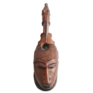 Puede incluir: Una escultura de madera de un rostro humano con tallas intrincadas. La escultura es de color marrón y tiene un cuello largo y delgado.