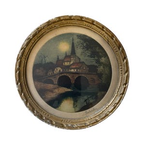 Puede incluir: Una impresi&oacute;n enmarcada de un paisaje nocturno con una iglesia, un puente y agua. El marco redondo es dorado con un dise&ntilde;o ornamentado. La obra de arte tiene un formato circular. La impresi&oacute;n muestra una luna llena.