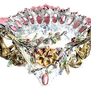 May include: A ceramic wall vase with a scalloped edge and two snake-like handles. The vase is decorated with flowers and leaves in shades of pink, yellow, green, and blue.