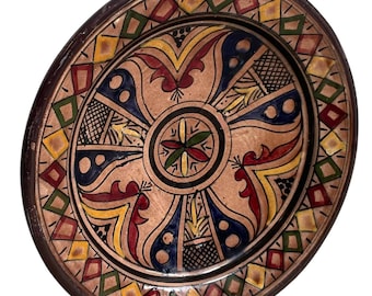 May include: A round, hand-painted ceramic plate with a dark brown rim. The plate features a central floral design and geometric patterns in red, yellow, green, and blue. The plate's diameter is approximately 10 inches.