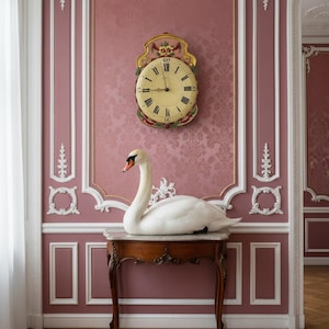 May include: A white swan with an orange beak rests on a small, ornate wooden table with a marble top. The table is set against a pink wall with decorative molding and a vintage clock. The clock has a cream-coloured face and black Roman numerals.