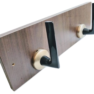 Puede incluir: Un perchero de pared de madera con dos ganchos de metal negro. Los ganchos están unidos a la madera con anillos de metal dorado.