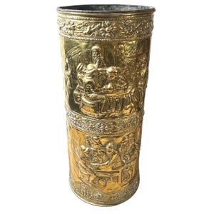 Könnte beinhalten: Ein hoher, zylindrischer Regenschirmständer aus Messing mit aufwendigen Reliefschnitzereien. Der Ständer zeigt detaillierte Szenen von Menschen, möglicherweise aus einem historischen oder allegorischen Kontext, und Blumenmuster. Das Messing hat eine polierte, goldene Oberfläche.