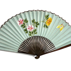 Puede incluir: Un abanico de mano azul claro con un diseño pintado de flores y una mariposa. El abanico está hecho de papel y tiene un mango de madera.