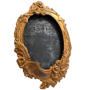 May include: A gold-toned oval picture frame with ornate details and a black background. The frame has a distressed finish and is made of metal.