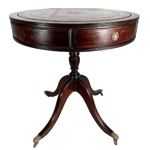 May include: A round, mahogany wood table with a leather top and a single drawer. The table has a pedestal base with three legs that end in brass paw feet.