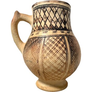 Może przedstawiać: Brązowy dzban ceramiczny z uchwytem i geometrycznym wzorem w czarnym kolorze. Wzór jest połączeniem rombów i kwadratów.