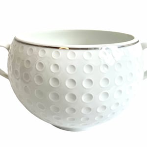 Op de afbeelding: Witte keramische soepkom met een gepunte oppervlakte die lijkt op een golfbal. De kom heeft een zilveren rand en twee handvatten.