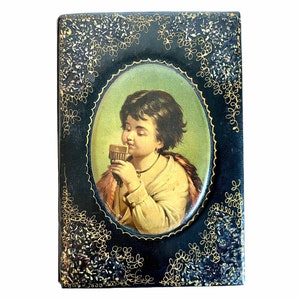 May include: A vintage black box with gold accents and a painted image of a child drinking from a cup. The box has a decorative border of gold swirls and flowers.