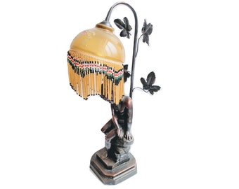 Könnte beinhalten: Eine Vintage-Tischlampe mit einem bronzenen Sockel, der eine sitzende Figur zeigt. Der Lampenschirm ist aus hellem gelbem Glas mit einem Perlenbesatz.