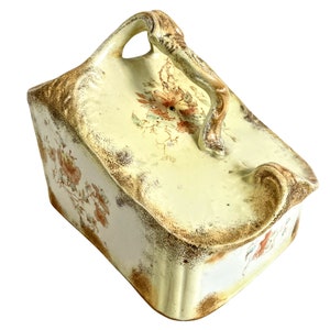 May include: A vintage ceramic box with a floral design and a handle. The box is yellow with brown and orange floral accents.