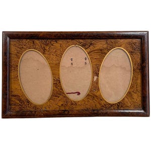 May include: A vintage wooden picture frame with three oval openings. The frame has a dark brown border and a patterned, light brown background. The oval openings have a gold-colored trim and are empty.
