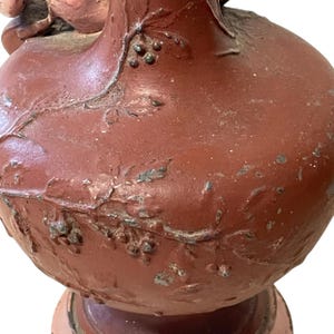 Pu&ograve; includere: Primo piano di un vaso in ceramica marrone-rossastro con superficie strutturata. Il vaso presenta disegni a rilievo di foglie e bacche. La vernice &egrave; usurata, rivelando indizi del materiale sottostante. Il vaso poggia su una base decorativa.