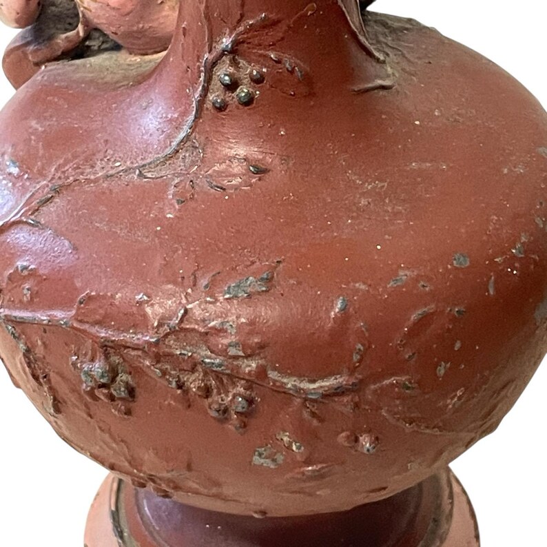 Pu&ograve; includere: Primo piano di un vaso in ceramica marrone-rossastro con superficie strutturata. Il vaso presenta disegni a rilievo di foglie e bacche. La vernice &egrave; usurata, rivelando indizi del materiale sottostante. Il vaso poggia su una base decorativa.