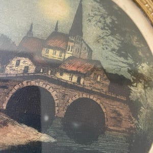 Puede incluir: Una impresi&oacute;n enmarcada de una escena de un pueblo europeo. La obra de arte representa un puente de piedra con aberturas arqueadas, que conduce a edificios y una iglesia con un alto campanario. La paleta de colores incluye marrones, rojos y verdes, creando una est&eacute;tica vintage.