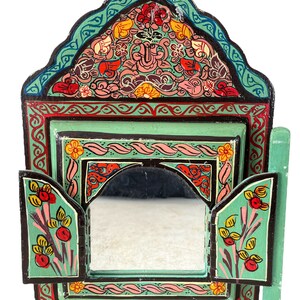Puede incluir: Un pequeño espejo de madera pintado de forma intrincada con un fondo verde y diseños florales en rosa, rojo, amarillo y azul. El espejo tiene una puerta con bisagras a cada lado.