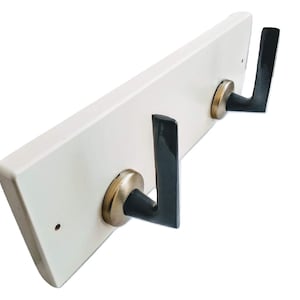 Puede incluir: Perchero de pared de madera blanca con dos ganchos de metal negro. Los ganchos tienen una base redondeada y un diseño en forma de V.