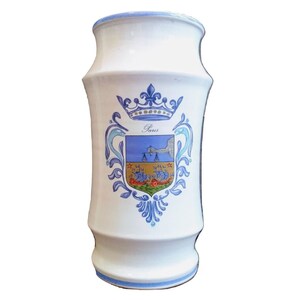 May include: White ceramic vase with a blue and gold design featuring the coat of arms of Paris, France. The vase has a cylindrical shape with a slightly flared top and bottom.