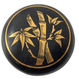 May include: A round, black decorative box with a gold bamboo design. The bamboo is depicted with detailed lines and leaf patterns, set against a black background. The box has a gold border.