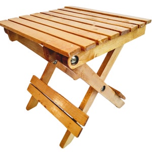 May include: A small, folding wooden table with a natural finish. The tabletop is made of parallel wooden slats. The legs are designed in a cross-legged style, allowing the table to fold flat for storage. The table is approximately 18 inches long, 12 inches wide, and 15 inches high.