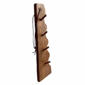 Op de afbeelding: Een houten wandrek met vijf haken, elk met een kleine houten pin. Het rek is gemaakt van donkerbruin hout en heeft een rustieke, handgemaakte uitstraling. Het is bevestigd aan de muur met een leren riem.