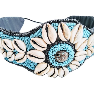 May include: A black beaded headband with a large blue and white shell design. The headband is decorated with cowrie shells and turquoise beads.