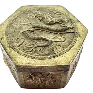 Puede incluir: Caja metálica hexagonal vintage con un relieve detallado de dragón en la tapa. La caja tiene un acabado dorado antiguo deslustrado y tallas florales en los laterales. El dragón está rodeado de nubes.