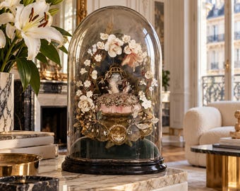 Op de afbeelding: Een glazen stolp met een decoratief arrangement van witte en crèmekleurige kunstbloemen, bladeren en een centraal kussen, geplaatst op een donkere ronde voet met een donkergroene fluwelen voering.
