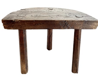 Puede incluir: Un taburete de madera rústico con una parte superior irregular y tres patas robustas. El taburete está hecho de madera marrón oscura con un acabado natural y desgastado.