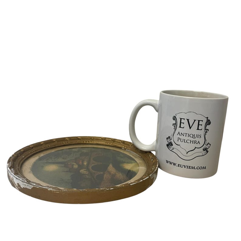 Puede incluir: Una taza de cer&aacute;mica blanca con asa y el texto "EVE ANTIQUIS PULCHRA" y una direcci&oacute;n web. Junto a ella, una imagen redonda enmarcada con un marco dorado y una escena de paisaje.