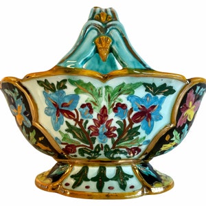 May include: A ceramic basket with a blue and green handle and a white base decorated with colorful floral patterns. The basket has a gold trim and is on a black and green base.