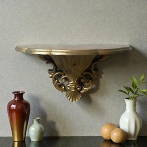 May include: A gold-colored wall shelf with ornate, classical detailing. The shelf is mounted on a gray wall, with decorative vases and spheres on a black surface below. The shelf has a semi-circular shape.