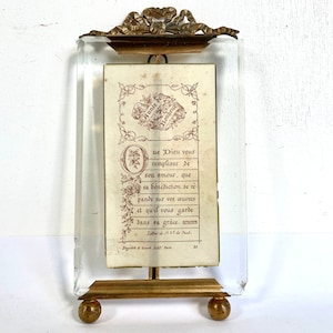 May include: A gold-toned metal frame with a glass front and a stand. The frame holds a piece of paper with a handwritten message in French. The message reads: "Que Dieu vous remplisse de son amour, que sa benediction se repande sur vos oeuvres et qu'il vous garde dans sa grace."