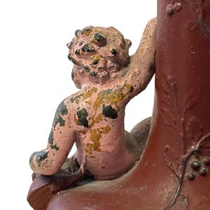 Pu&ograve; includere: Vista dettagliata di un vaso rosso antico decorativo con una figura di cherubino. Il cherubino &egrave; dipinto in rosa e oro, con vernice scrostata. Il vaso ha una superficie strutturata ed &egrave; adornato da motivi a foglie e uva.