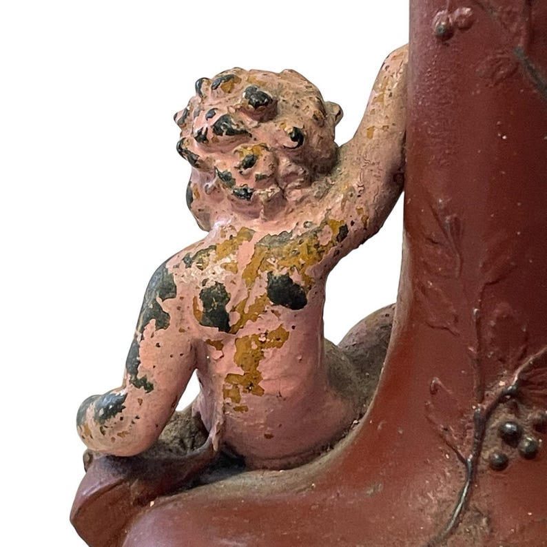 Pu&ograve; includere: Vista dettagliata di un vaso rosso antico decorativo con una figura di cherubino. Il cherubino &egrave; dipinto in rosa e oro, con vernice scrostata. Il vaso ha una superficie strutturata ed &egrave; adornato da motivi a foglie e uva.