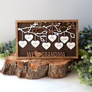 May include: Wooden sign with a tree branch design and hearts hanging from the branches. Each heart has a name written on it: Audrey, William, Nathan, Sarah, Abby, Elizabeth, Jacob, and Hunter. The sign reads "We ♥ Grandma".