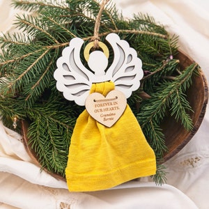 May include: White wooden angel ornament with a yellow fabric pouch and a heart-shaped wooden tag that reads "Forever in our hearts, Grandma Bernie".