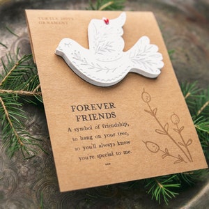 Może przedstawiać: Biała ozdoba w kształcie turkawki z wygrawerowanymi wzorami liści, przymocowana do prostokątnej brązowej karty. Karta zawiera napis TURTLE DOVE ORNAMENT, FOREVER FRIENDS, wiersz o przyjaźni oraz rysunek liniowy rośliny z jagodami.