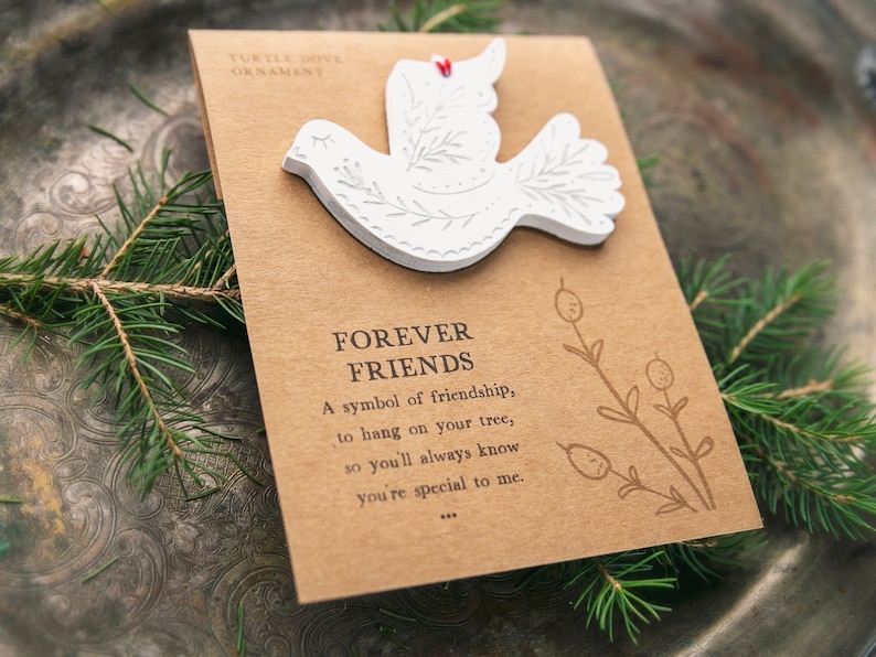 Może przedstawiać: Biała ozdoba w kształcie turkawki z wygrawerowanymi wzorami liści, przymocowana do prostokątnej brązowej karty. Karta zawiera napis TURTLE DOVE ORNAMENT, FOREVER FRIENDS, wiersz o przyjaźni oraz rysunek liniowy rośliny z jagodami.