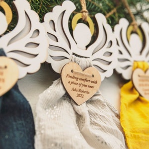 May include: Three white wooden angel ornaments with wings and a heart-shaped tag. Each tag has a different message. The first tag says "Finding comfort with a piece of you near". The second tag says "Finding comfort with a piece of you near Ada Roberts 2022". The third tag says "Finding comfort with a piece of you near Ada Roberts 2022".