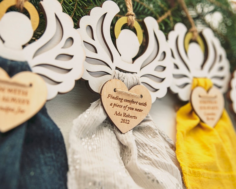 May include: Three white wooden angel ornaments with wings and a heart-shaped tag. Each tag has a different message. The first tag says "Finding comfort with a piece of you near". The second tag says "Finding comfort with a piece of you near Ada Roberts 2022". The third tag says "Finding comfort with a piece of you near Ada Roberts 2022".