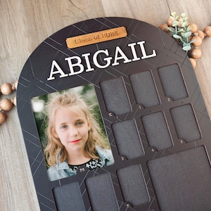 May include: Black wooden photo frame with a white arch and the text "Class of 2024" and "ABIGAIL". The frame has 12 photo slots and a larger photo slot at the top. A photo of a young girl is in the larger slot.