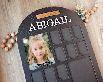 May include: Black wooden photo frame with a white arch and the text "Class of 2024" and "ABIGAIL". The frame has 12 photo slots and a larger photo slot at the top. A photo of a young girl is in the larger slot.