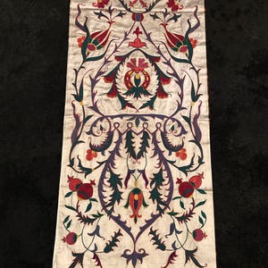 May include: A white fabric panel with a traditional floral design embroidered in shades of red, green, purple, and gold. The design features stylized flowers, leaves, and pomegranates.