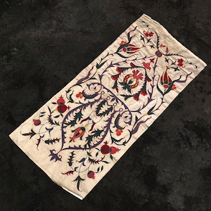 May include: A white fabric panel with a floral design embroidered in shades of red, green, purple, and brown. The design features stylized flowers, leaves, and pomegranates.
