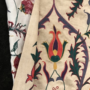 May include: Close-up of a cream-colored fabric with intricate embroidery. The design features stylized flowers in shades of red, orange, yellow, green, and purple.