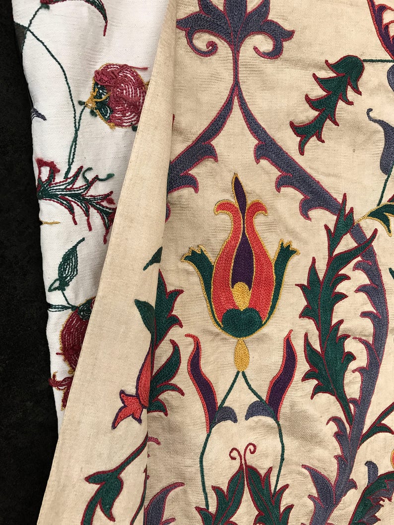 May include: Close-up of a cream-colored fabric with intricate embroidery. The design features stylized flowers in shades of red, orange, yellow, green, and purple.
