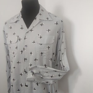 Peut inclure: Une chemise à manches longues grise avec un motif de croix noir et blanc. La chemise a un col et deux poches.